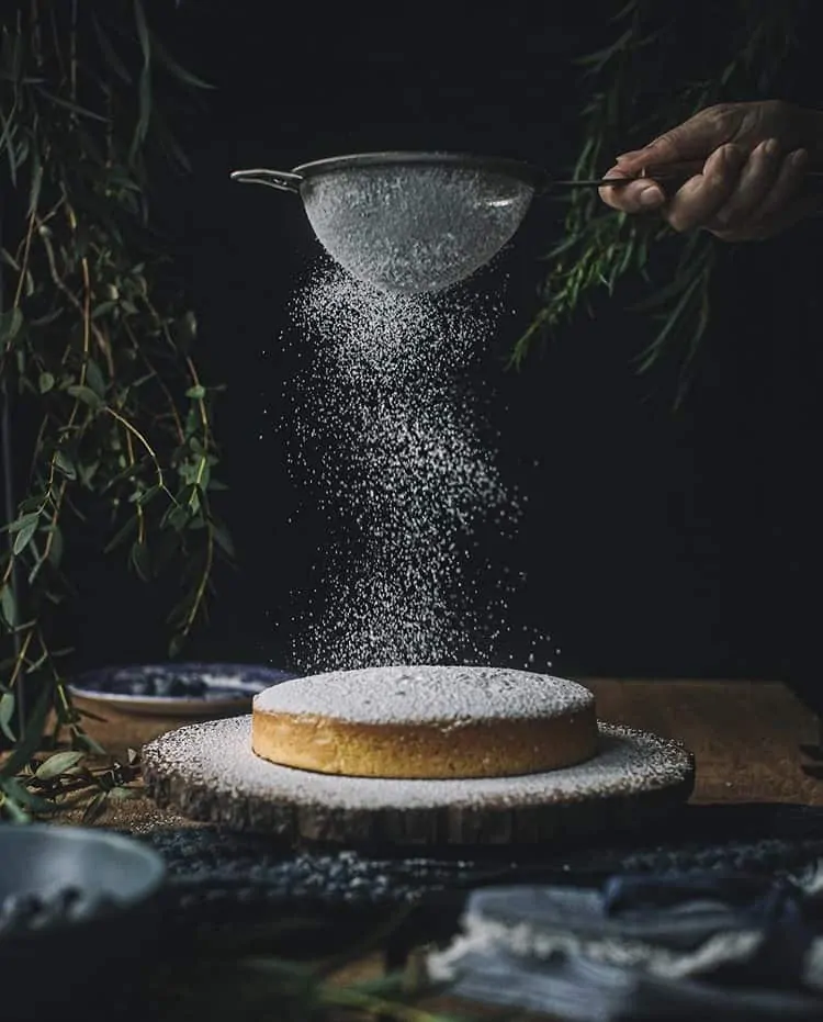 one layer of cake is set on a back background. a hand is sifting powdered sugar over cake