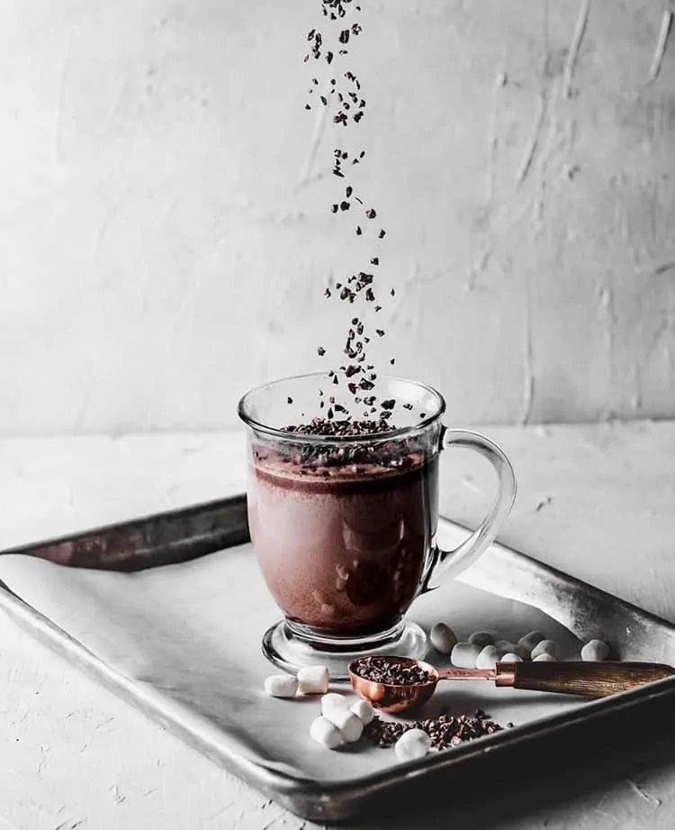 clear mug with hot chocolate on top of a black baking tray with a layer of parchment. chocolate sprinkles are suspended in midair