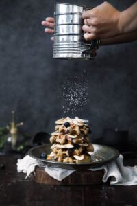 stack of waffles with blueberries on a wooden slab. a pair of hands a sifting powdered sugar over the top